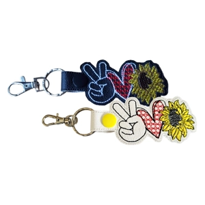 Two Peace Love Sunflower keyrings shown together, one in black and one in white, each featuring an embroidered peace sign hand, stitched heart and sunflower, finished with a metal ring and lobster clasp.