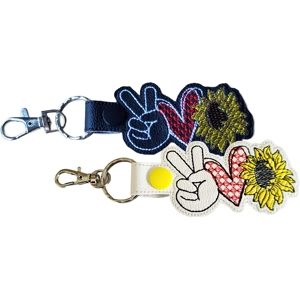 Two embroidered keyrings on a white background: one in black and one in white. Each keyring features a peace hand sign, a red heart, and a yellow sunflower stitched onto faux leather, attached to a metal clip and key ring.