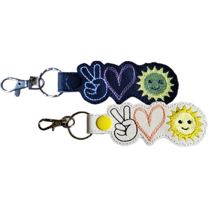 Two embroidered snap-tab keyrings, each shaped around three stitched icons: a peace-sign hand, a heart and a smiling sun. One keyring is made from black faux leather with white, pink and yellow stitching. The other is made from white faux leather with black, orange and yellow stitching. Both keyrings have silver hardware.