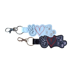 Two embroidered snap-tab keyrings, each shaped around three stitched icons: a peace-sign hand, a heart and a teddy bear. One keyring is made from white faux leather with black, red and light brown stitching. The other is made from black faux leather with white, red and light purple stitching. Both keyrings have silver hardware.