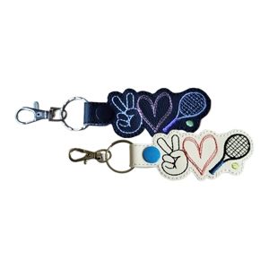 Two embroidered snap-tab keyrings, each shaped around three stitched icons: a peace-sign hand, a heart and a tennis racket with a tennis ball. One keyring is made from black faux leather with white, pink, grey and purple stitching. The other is made from white faux leather with black, red, blue and black stitching. Both keyrings have silver hardware.