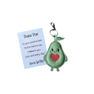 A handmade pocket pal keyring featuring a smiling green felt pear with stitched eyes, a red heart on its belly, and a small felt leaf on top. Attached is a card with an encouraging poem titled ‘Positive Pear’.