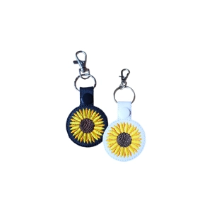 Two round embroidered keyrings featuring bright yellow sunflowers with brown centres. One keyring is made of black faux leather with a black snap fastener, and the other is white faux leather with a white snap fastener. Both have silver key rings and lobster-claw clasps attached at the top.