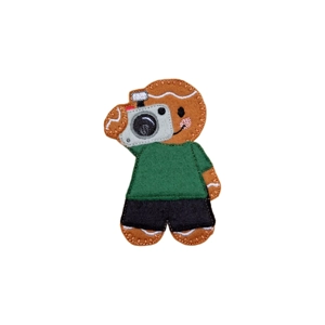 A felt gingerbread photographer character holding a stitched camera, wearing a green top and black trousers, with embroidered details.