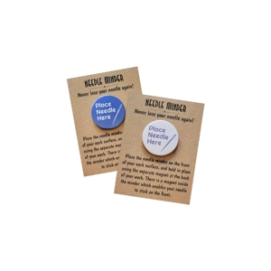 Two round 38 mm needle minders with the phrase “Place Needle Here” and a needle illustration, shown in blue and white designs on kraft backing cards for sewing and embroidery projects.