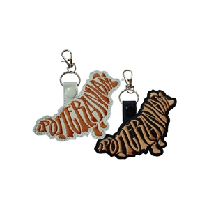 Two Pomeranian-shaped keyrings made from faux leather, one with a white base and one with a black base. Each keyring features embroidered word-art forming the silhouette of a Pomeranian dog, with metal clips attached for fastening to keys or bags.