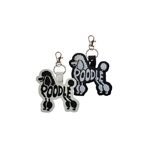 Two Poodle-shaped faux leather keyrings, one white with black embroidery and one black with silver embroidery. Each keyring features a stitched Poodle silhouette with the word ‘POODLE’ integrated into the design, and both have a snap tab, keyring and metal clasp attached.