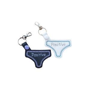 Two Positive Pants keyrings, one made from black faux leather and one from white faux leather, each embroidered with the word ‘Positive’ and finished with a metal keyring and lobster clasp.