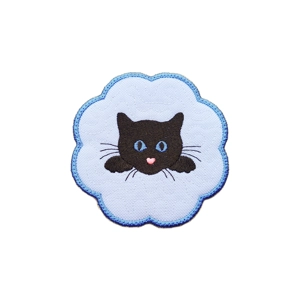 A scalloped white fabric candle coaster with a blue stitched border, featuring an embroidered black cat peeking over the edge with bright blue eyes, fine whiskers and a small pink nose.