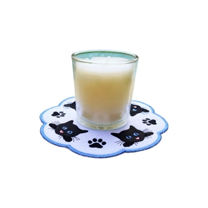 A glass candle placed on a scalloped fabric candle mat with a blue stitched border and contrasting embroidered cat faces and paw prints arranged in a circular pattern.