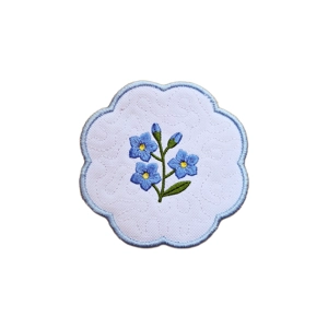 A scalloped fabric candle coaster with embroidered blue forget me not flowers, green stems and leaves, a soft stippled texture, and a pale blue stitched border.