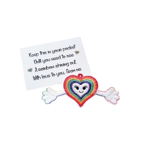 A rainbow-striped heart-shaped felt pocket hug keyring with a smiling face and white outstretched arms ending in mitten-shaped hands. It is placed in front of a keepsake card that reads: ‘Keep this in your pocket until you need to see a rainbow shining out, with love to you, from me’. Small rainbow heart icons decorate the card.
