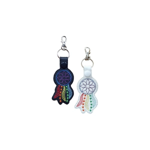 Two faux-leather dream catcher keyrings, one with a black base and one with a white base. Both feature embroidered circular dream catcher designs with rainbow-coloured hanging feather details. Each keyring has a snap tab at the top and a metal keyring with a lobster clasp.