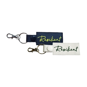 Two faux leather keyrings with the word 'Resilient embroidered on them. One keyring is black with yellow embroidery and a black snap fastener, attached to a silver keyring and silver lobster clasp. The other is white with dark green embroidery, a white snap fastener, and a silver keyring with a silver lobster clasp. Both keyrings are rectangular with textured stitching.