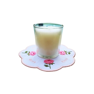 White candle in a clear glass holder placed on a round embroidered mat with pink roses and the word “Rose” stitched around the scalloped edge.