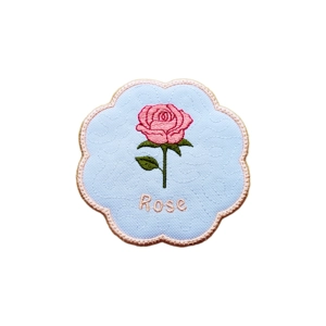 Handmade embroidered Rose Coaster Mat – June Birth Flower. Features a pink rose with green leaves stitched on a white scalloped fabric mat with a pale pink border and the word Rose embroidered beneath the flower.
