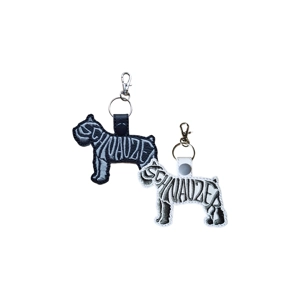 Two Schnauzer-shaped faux leather keyrings with embroidered word-art reading 'Schnauzer'. One keyring is black with grey stitching, and the other is white with grey stitching. Both have snap-closure tabs and metal clasps for attaching to keys or bags.