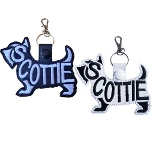 Two Scottie word art keyrings shown in black faux leather with white stitching and white faux leather with black stitching, both finished with metal clasp hardware.