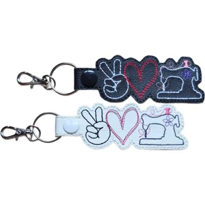 Two Sewing Peace Love keyrings in black and white faux leather, each featuring a peace sign hand, stitched heart and sewing machine design with sturdy metal ring and lobster clasp.