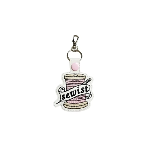 Sewist keyring made of white faux leather featuring an embroidered pink thread spool with a needle and a banner reading sewist, attached to a pink snap strap with a metal keyring and lobster clasp.