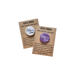 Two round needle minders on brown kraft backing cards reading 'NEEDLE MINDER – Never lose your needle again!'. One minder is white with navy text reading 'Sew Sew' with a needle graphic forming a line between the words, and the other is purple with white text reading 'Sew Sew' with the same needle graphic. Both cards include instructions for using the magnetic needle minders for sewing or embroidery.