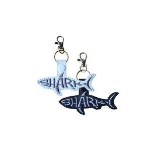 Two shark word art keyrings, one in black with light stitching and one in white with dark stitching, with the word “Shark” stitched to form the shape of a shark, each finished with a metal clasp and keyring.