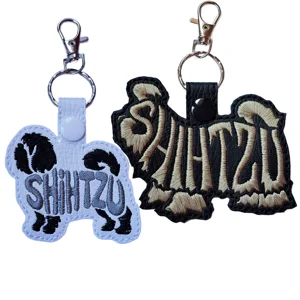 Two Shih Tzu word art keyrings shown in white faux leather with black and grey stitching and black faux leather with tan stitching, both finished with metal clasp hardware.
