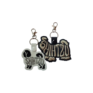 Two Shih Tzu silhouette word-art keyrings shown side by side, one in white faux leather and one in black, each machine embroidered with the word “Shih Tzu,” finished with a metal keyring and clasp.