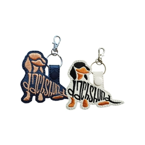 Two embroidered sitting Dachshund silhouette keyrings shown on a white background, one in black and one in white. Each features stitched lettering forming the word Dachshund, with a metal lobster clasp and keyring attached.