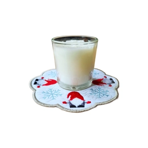 A white candle in a clear glass holder sits on a scalloped embroidered table mat decorated with festive gnomes wearing red hats and surrounded by blue snowflakes.