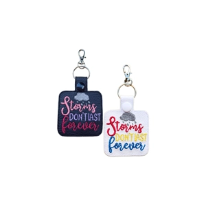 Two embroidered keyrings shown side by side. One has a black base with pink and purple stitching, and the other has a white base with red, yellow and blue stitching. Both feature a small grey cloud with raindrops and the phrase 'Storms Don't Last Forever' in colourful text, each attached to a metal keyring and clasp.