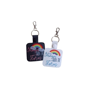 Two embroidered keyrings shown side by side. One has a black base with rainbow stitching and the phrase 'Stronger Than the Storm' in pink and blue thread, and the other has a white base with the same rainbow and cloud design stitched in blue thread. Both keyrings are attached to metal keyrings and lobster clasps.