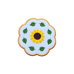 A round embroidered candle coaster featuring a central yellow sunflower with a dark centre, surrounded by green leaves on a white background, finished with a scalloped stitched border.