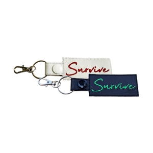 Two faux-leather keyrings embroidered with the word Survive. One keyring is white with red stitching and a white snap fastener. The other keyring is black with green stitching and a black snap fastener. Both keyrings are fitted with silver-tone lobster clasps and split rings.