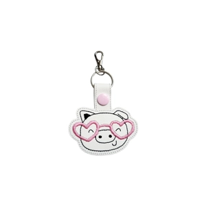 A small faux leather keyring featuring an embroidered smiling pig wearing pink heart-shaped glasses, with a pink snap fastener tab and silver keyring and lobster clasp.
