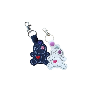 Two faux leather voodoo doll keyrings, one black and one white, each with stitched details including a heart, button-style eye, and tuft of hair, finished with a metal clasp and keyring.