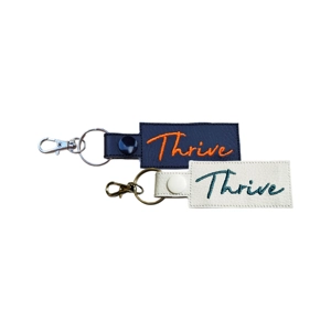 Black and white faux leather keyrings featuring the word Thrive embroidered in orange on the black keyring and teal on the white keyring. Both keyrings have silver-tone clasps and split rings.