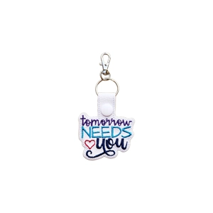 A white faux leather keyring embroidered with the words ‘tomorrow NEEDS you’ in purple, turquoise and navy blue thread, with a small red heart. The keyring has a white snap fastener, a silver split ring and a silver lobster clasp.