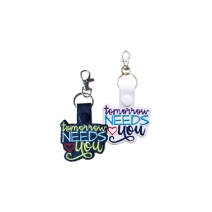 Two 'Tomorrow Needs You' keyrings, one black and one white, both made from faux leather and machine embroidered with colourful text and a small red heart, each with a metal keyring and clasp.