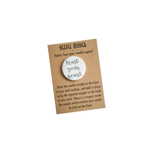 A round white needle minder with the words “Trust your heart” in soft grey lettering, accented with small heart details, displayed on a kraft needle minder backing card with printed instructions.