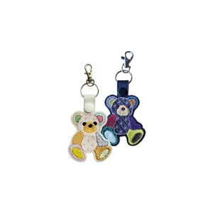 Two faux-leather keyrings featuring embroidered patchwork teddy bears. One keyring is white with multicoloured patchwork sections in yellow, green, pink, blue, and orange, finished with a white snap tab. The other keyring is black with brightly coloured patchwork sections in purple, blue, red, yellow, and green, finished with a black snap tab. Both include a metal keyring and a lobster clasp.