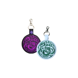 Two round faux-leather keyrings with embroidered dragon designs. One keyring has a black base with bright purple dragon stitching and a purple snap tab; the other has a white base with green dragon stitching and a white snap tab. Both have metal clasps at the top for attaching to bags or keys.