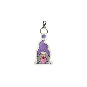 White faux leather keyring featuring an embroidered girl gnome with a tall purple hat, long grey braids, and a pink dress decorated with a small red flower. Finished with a purple snap and silver lobster clasp.