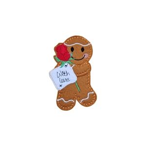 A felt gingerbread character with embroidered details, holding a red rose and a stitched ‘With Love’ tag, with a smiling face and rosy cheeks.
