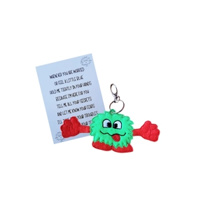 A cheerful felt worry monster pocket hug keyring with green body, red arms and feet, smiling face with tongue out, attached to a metal keyring clip and shown beside a printed keepsake card with a comforting poem.