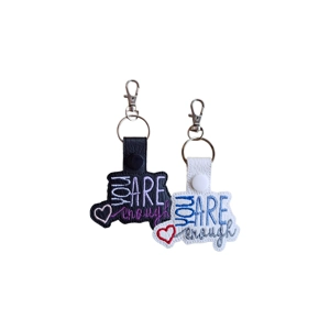 Two faux leather You Are Enough keyrings, one with a black backing and one with a white backing. Each features embroidered text with a small heart detail and a metal keyring attachment.