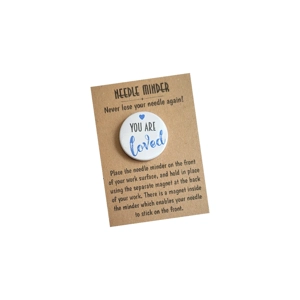 A round white needle minder with the words “You are loved” in soft lettering and a small heart detail, displayed on a kraft needle minder backing card with printed instructions.