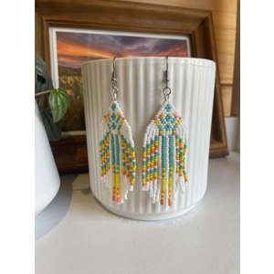 Handmade beaded earrings in shades of pastels and white, featuring an intricate geometric pattern and long fringe design. The earrings hang from silver hooks and are displayed against a textured white ceramic background