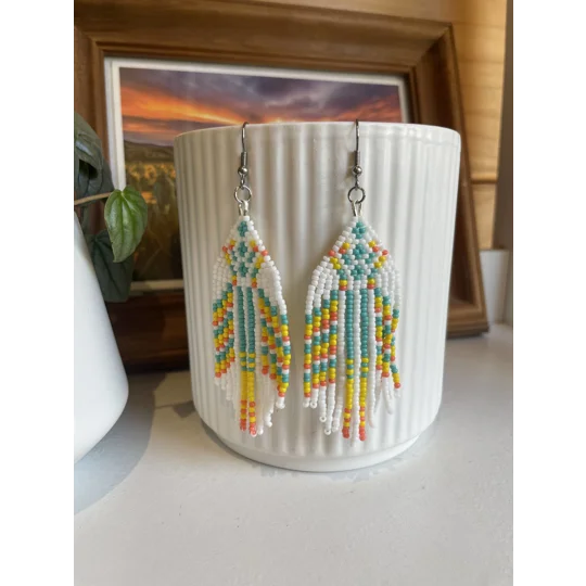 Handmade beaded earrings in shades of pastels and white, featuring an intricate geometric pattern and long fringe design. The earrings hang from silver hooks and are displayed against a textured white ceramic background