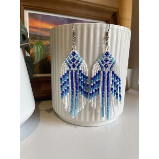 Handmade beaded earrings in shades of blue and white, featuring an intricate geometric pattern and long fringe design. The earrings hang from silver hooks and are displayed against a textured white ceramic background